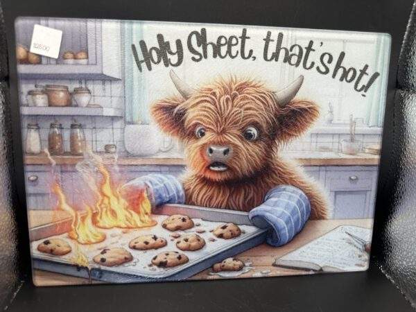 A cutting board with a cartoonish highland cow in a kitchen wearing oven mitts and holding a flaming batch of chocolate chip cookies on a cookie sheet. The text "Holy sheet, that's hot" is above its head.