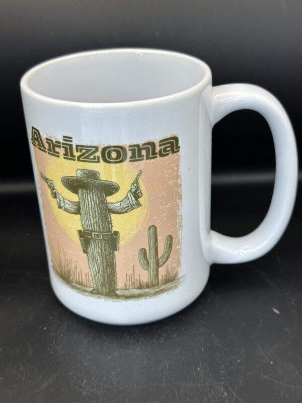 A white coffee mug with a square print on the front with the word "Arizona" over a gunslinging saguaro cactus with a cowboy hat, standing in front of the orange sky and yellow sun.