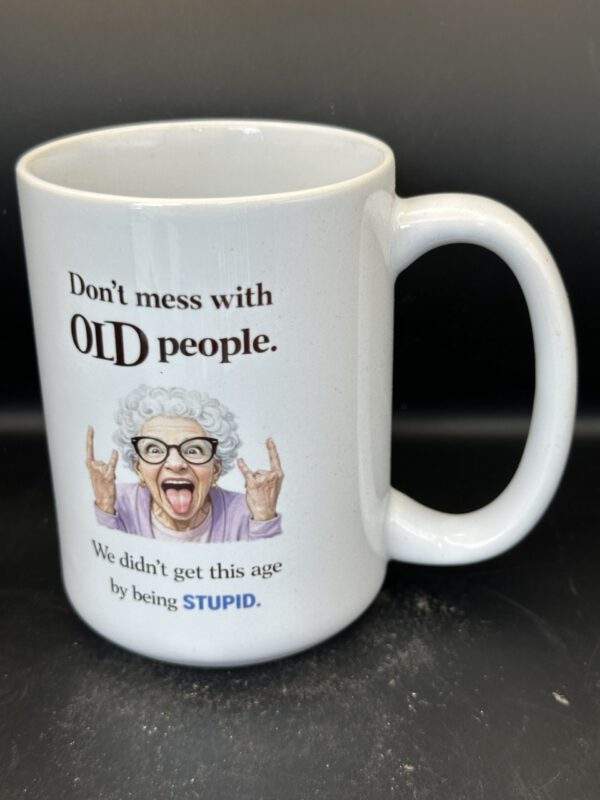 White mug with an elderly woman with horn-rimmed glasses sticking out her tongue and making rock and roll 'horns' hands. The text says, 'Don't mess with old people. We didn't get this age by being stupid.'