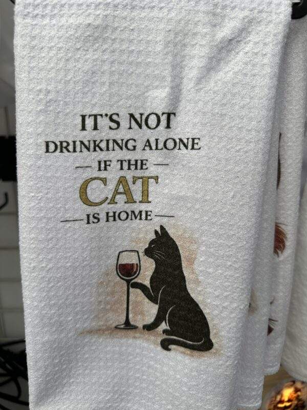 A white dish towel with an elegant black cat and a glass of wine. Text reads, 'It's not drinking alone if that cat is home'