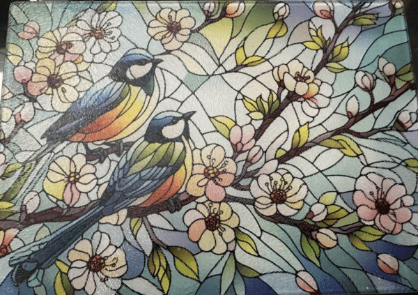 A glass cutting board with a stained-glass style design showing blue and yello songbirds on the branch of a flowering tree.