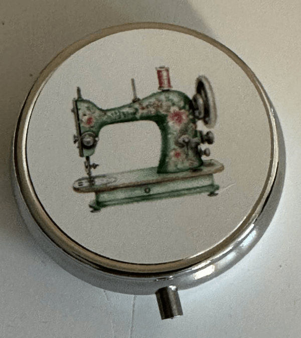 A round metal pill box with an image of a green sewing machine on the lid.