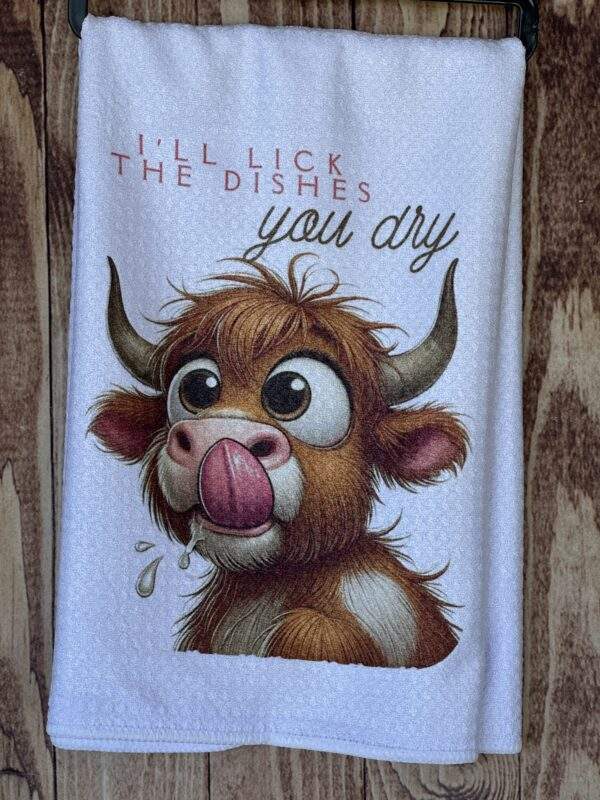 White, waffle weave kitchen dish towel with image of a funny cow licking his mouth. Text reads "I'll lick the dishes, you dry"