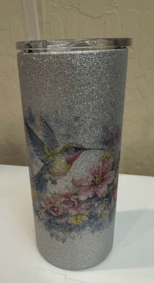 A glittering silver tumbler with lid and straw and design of a brilliant hummingbird and flowers.